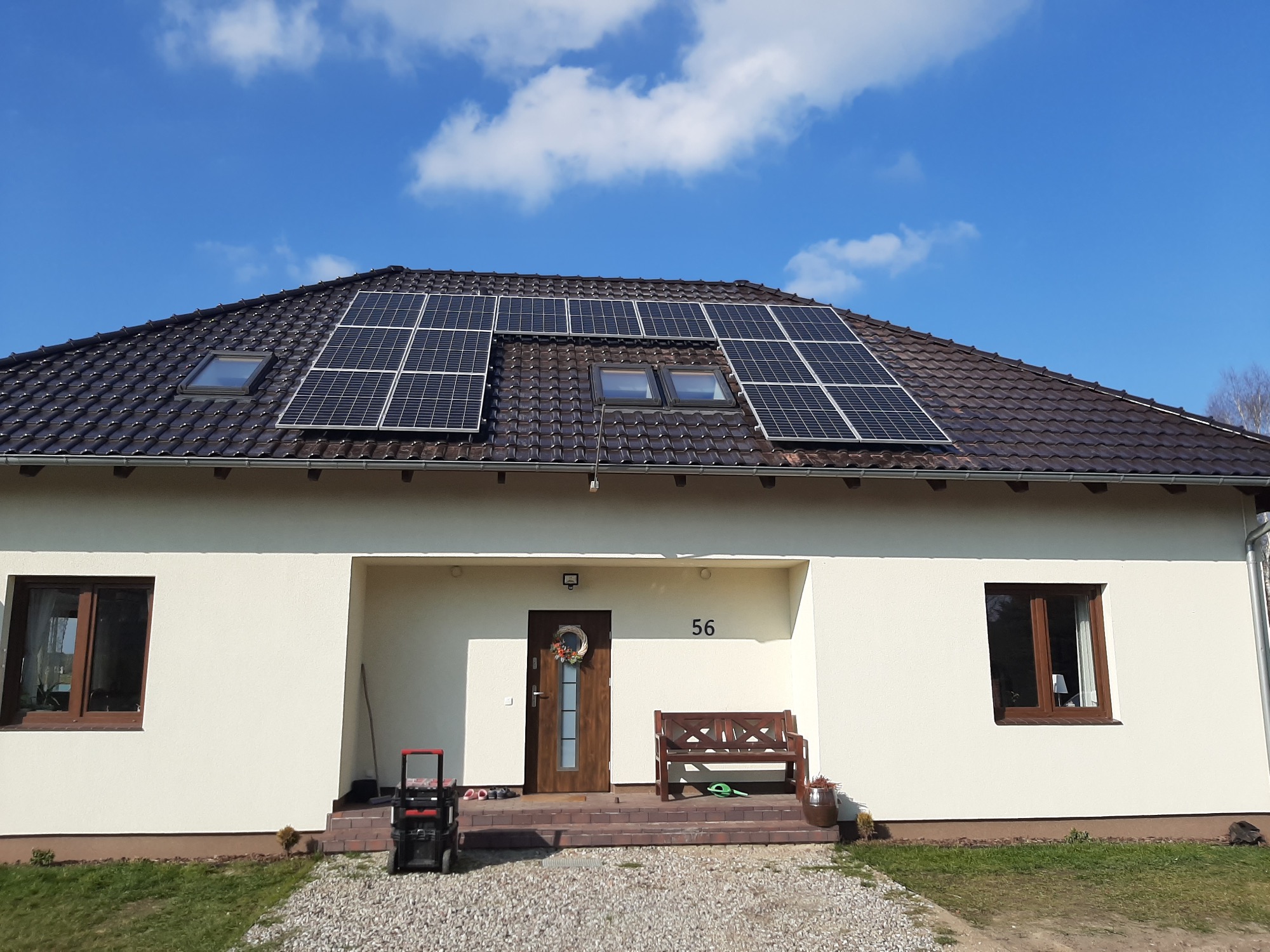 Dom jednorodzinny z brązowym dachem pokrytym panelami słonecznymi i dwoma oknami dachowymi, numer domu 56 nad wejściem, narzędzia na schodach.