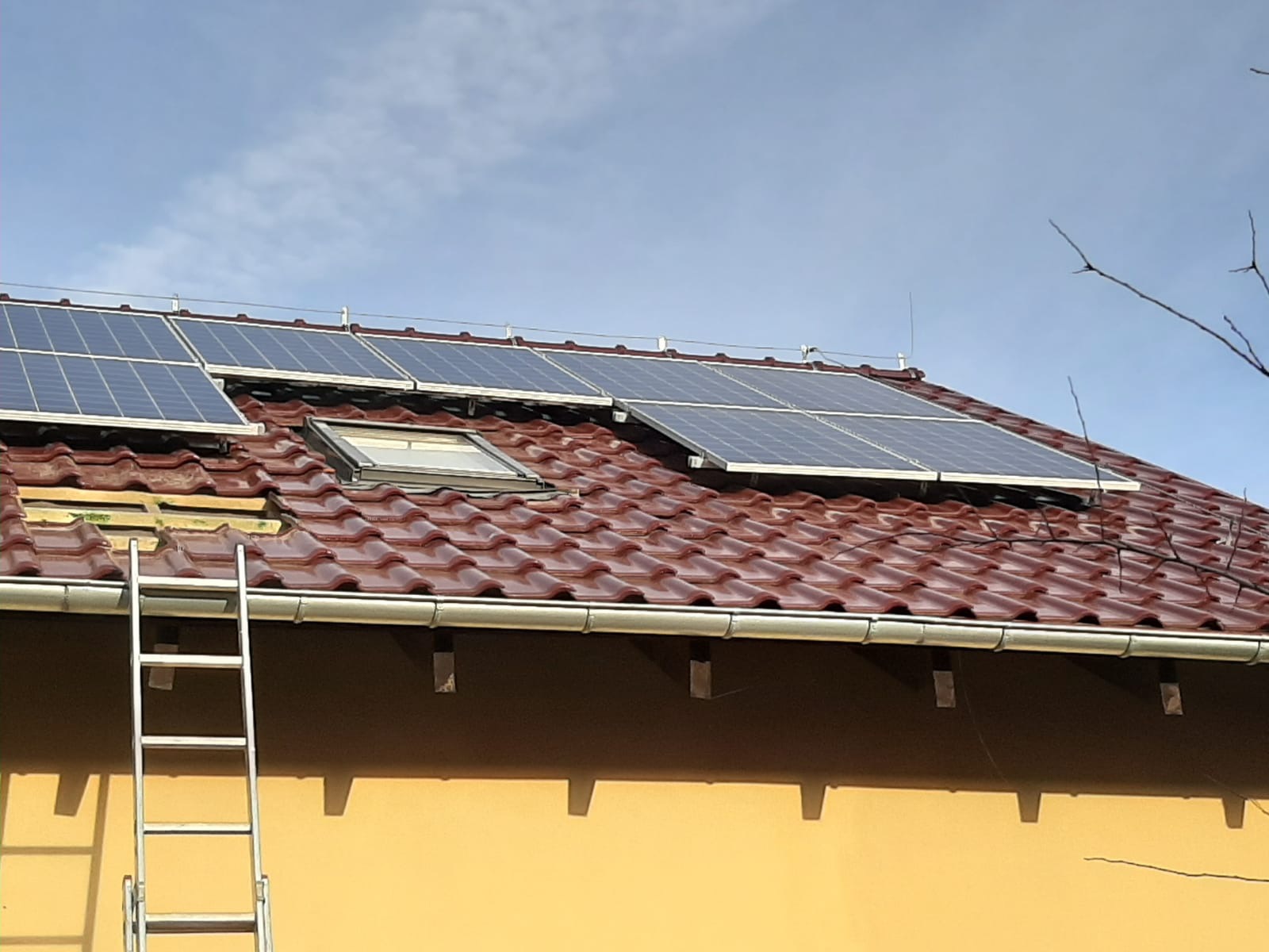 Montaż paneli słonecznych na dachu z czerwonej dachówki, widoczna drabina oparta o ścianę budynku.
