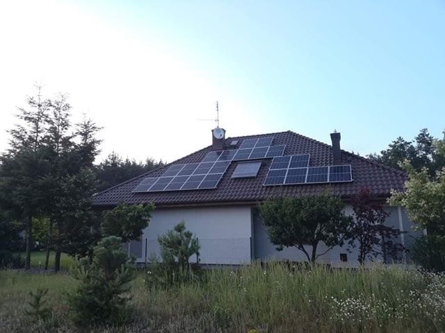 Dom jednorodzinny w Kostrzynie z instalacją fotowoltaiczną na brązowym dachu, antena satelitarna i komin widoczne, otoczony zielenią.