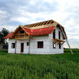 Dom w trakcie budowy z widocznym szkieletem dachu i czerwoną membraną dachową, zabezpieczony folią Tyvek, drabina oparta o ścianę, w tle pole zboża i pochmurne niebo.