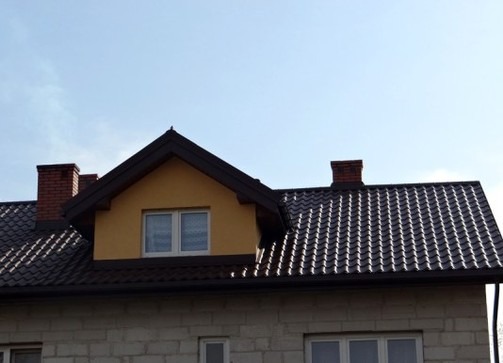 Dom z ciemnobrązowym dachem z dachówki, żółtą lukarną i dwoma kominami z cegły, sfotografowany w słoneczny dzień.