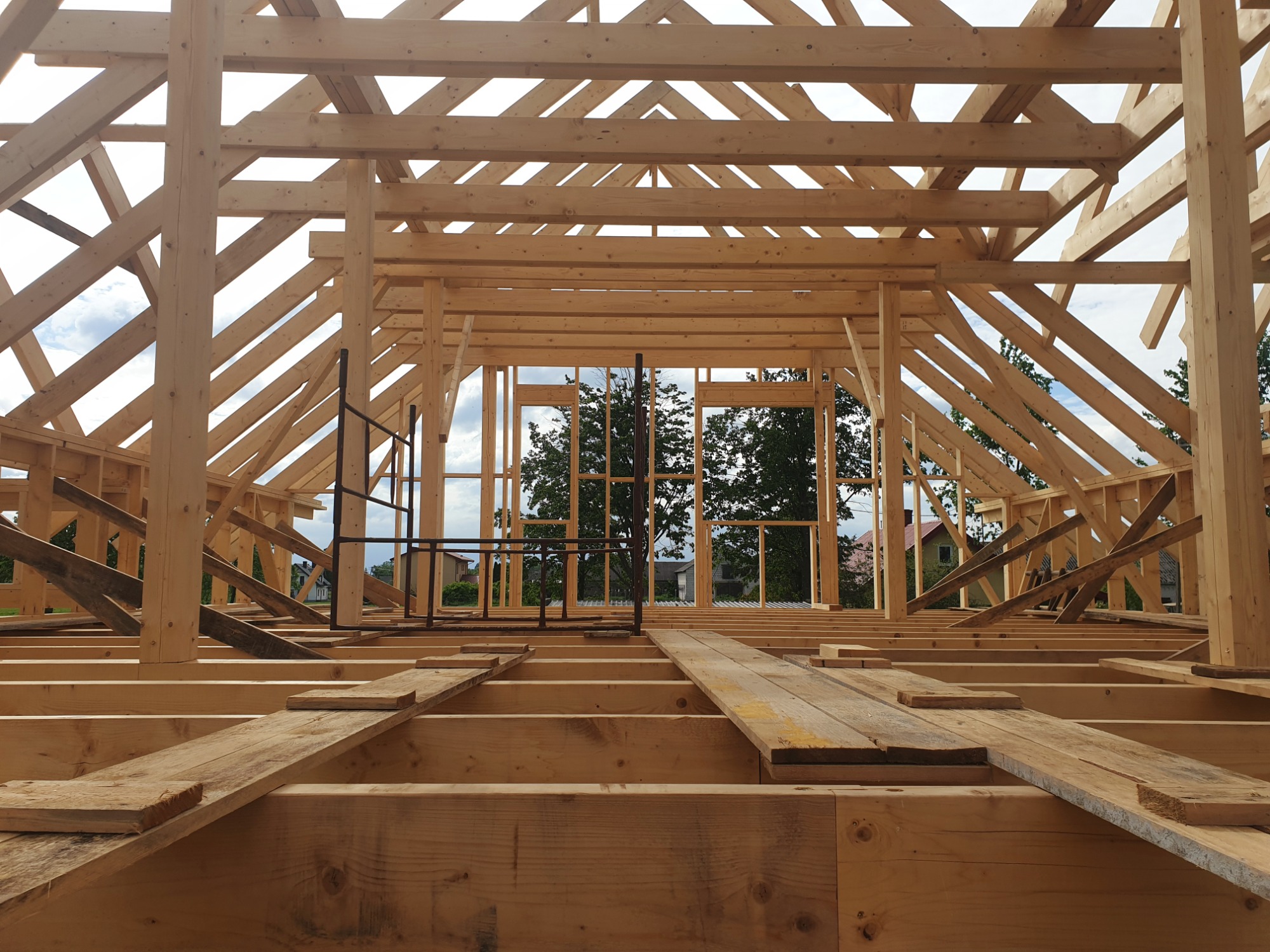 Konstrukcja drewniana dachu w trakcie budowy, widoczne elementy więźby dachowej z surowego drewna, na tle zieleni drzew i fragmentów zabudowy mieszkalnej.