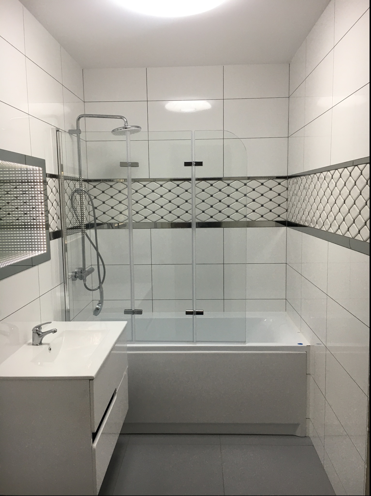 Nowoczesna łazienka po remoncie z białymi płytkami, dekoracyjnym pasem płytek w romby, kabiną nawannową i umywalką z szafką w minimalistycznym stylu.