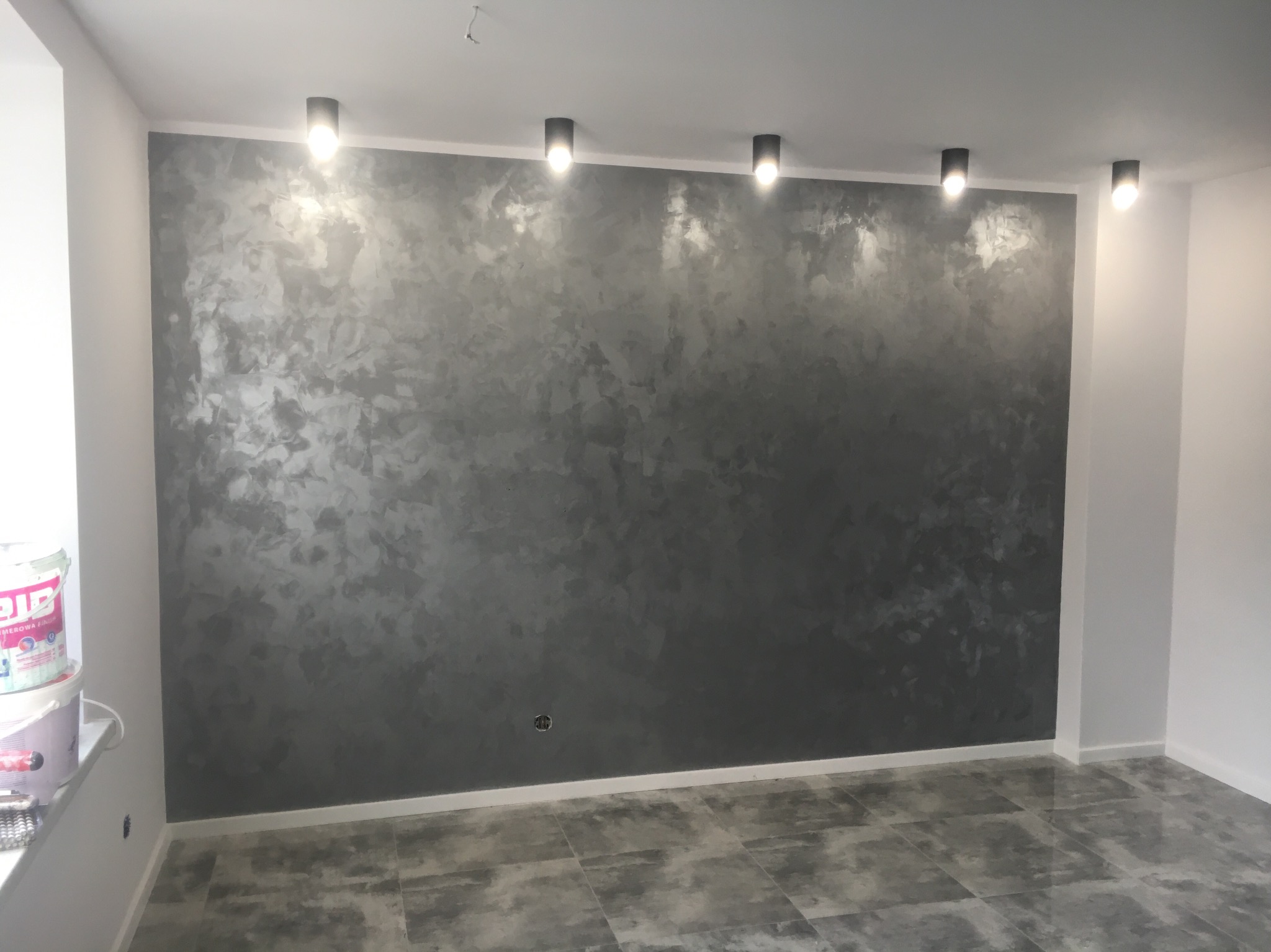 Ściana w pokoju po nałożeniu dekoracyjnej, szarej farby ze srebrnymi refleksami, oświetlona przez pięć punktowych lamp sufitowych. Widoczna puszka z farbą przy oknie.