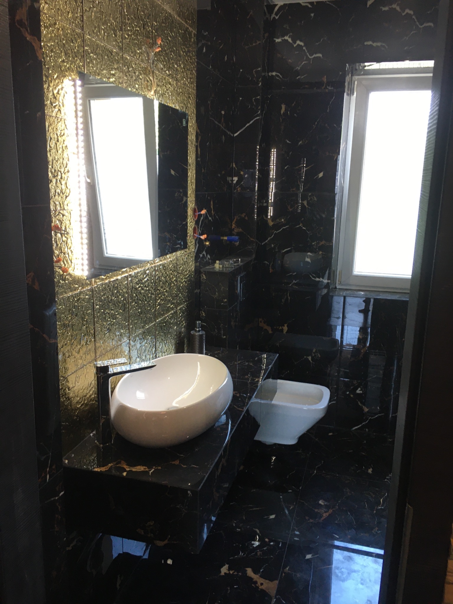 Elegancka łazienka w Jaworznie z czarnymi marmurowymi płytkami i złotą mozaiką, owalna umywalka na blacie, podświetlane lustro i bidet.