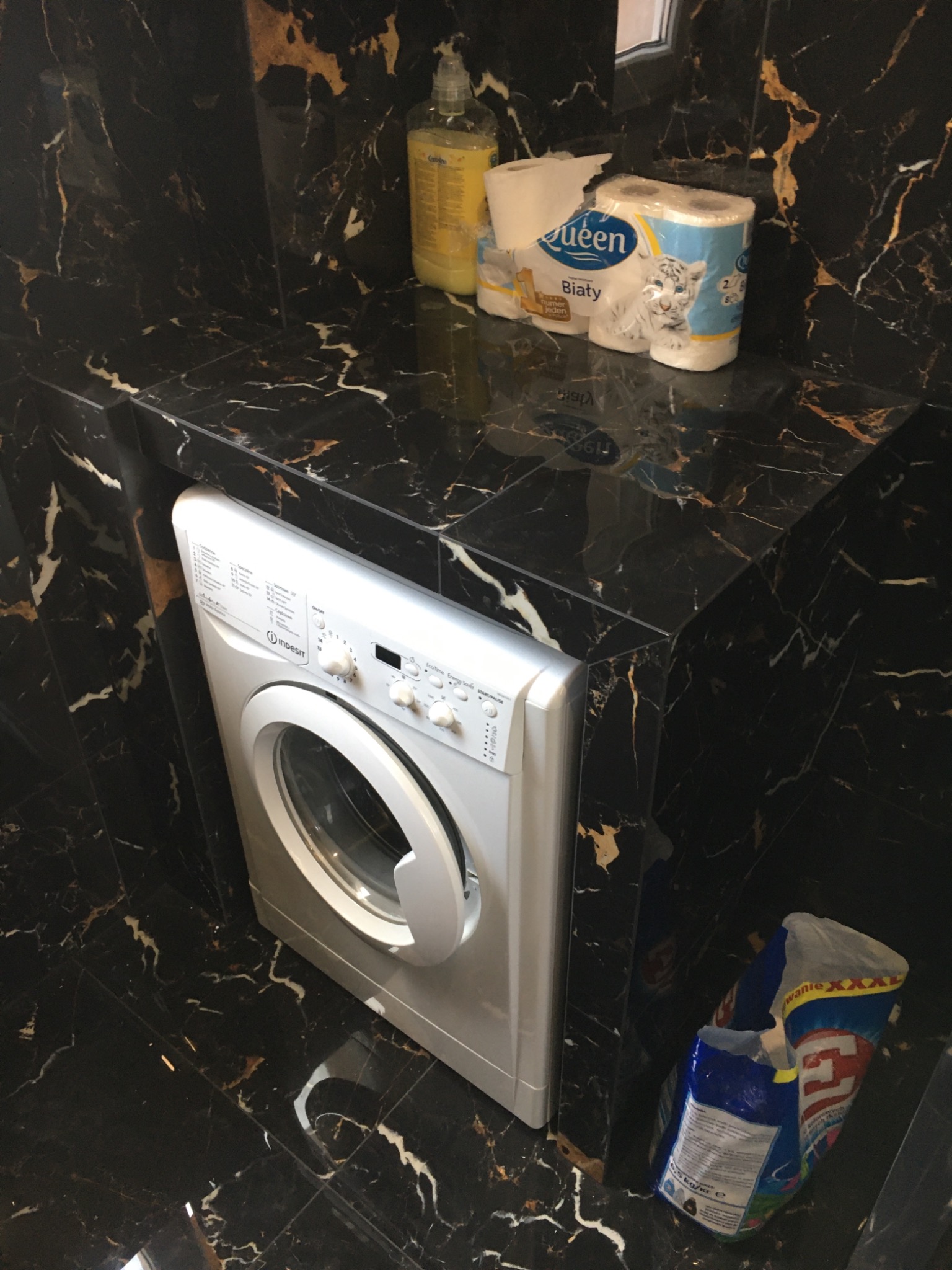 Biała pralka Indesit w łazience wykończonej czarnym marmurem z złotymi żyłami, obok środki czystości i proszek do prania.