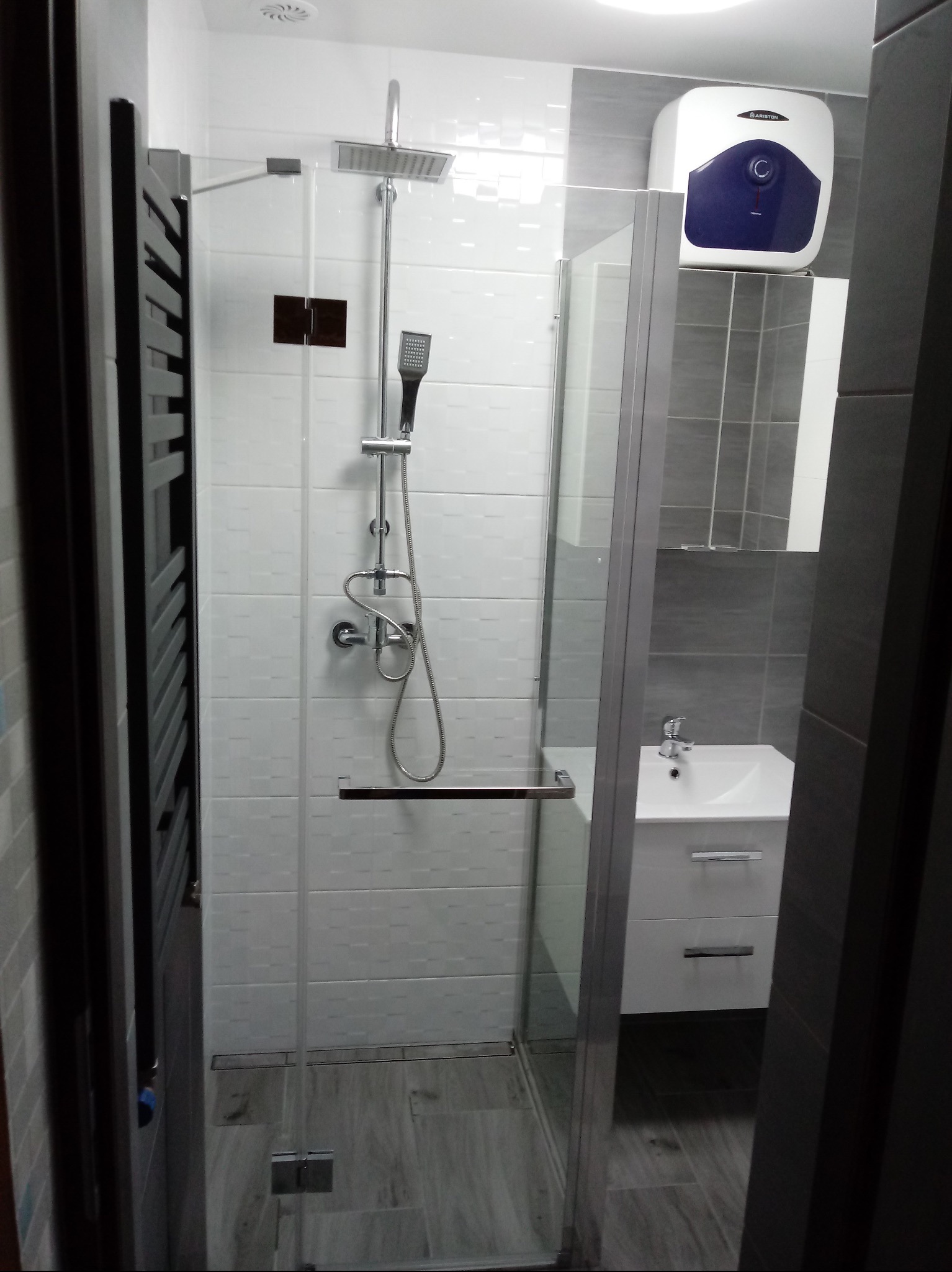 Nowoczesna kabina prysznicowa z deszczownicą i panelem prysznicowym, białe i szare płytki, grzejnik drabinkowy w łazience po remoncie, widoczny bojler elektryczny.