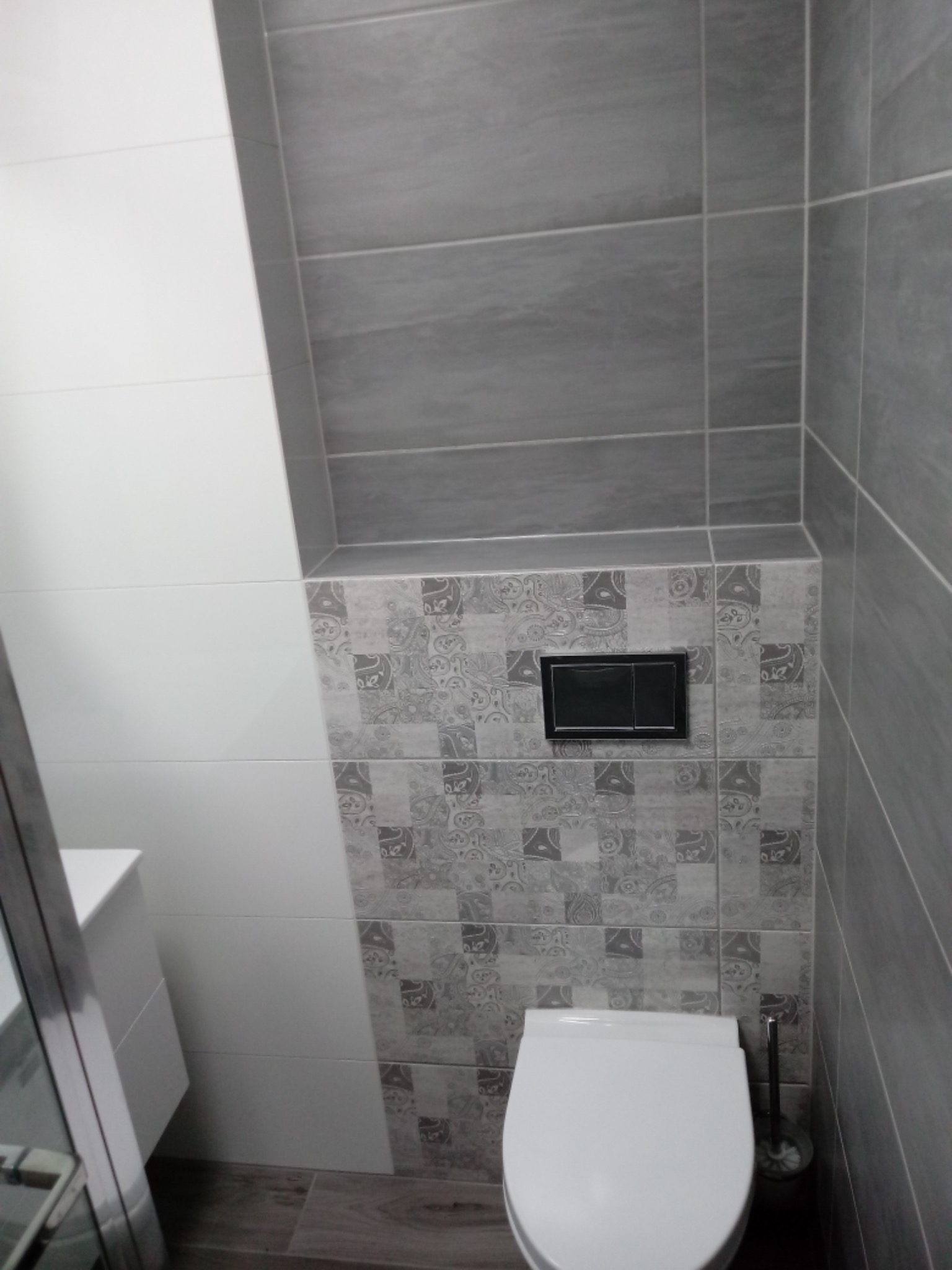 Nowoczesna toaleta z szarą i białą glazurą, wzorzyste płytki nad zabudowaną spłuczką, drewnopodobna podłoga.