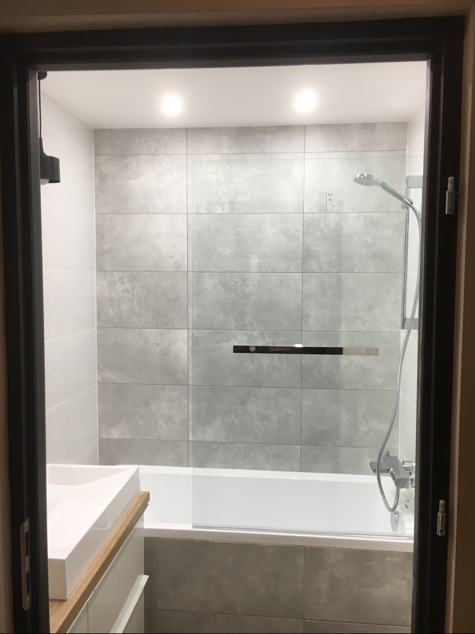 Nowoczesna łazienka z szarymi płytkami imitującymi beton, białą wanną z parawanem i umywalką nablatową na drewnianym blacie, widok zza ciemnej ramy drzwi.