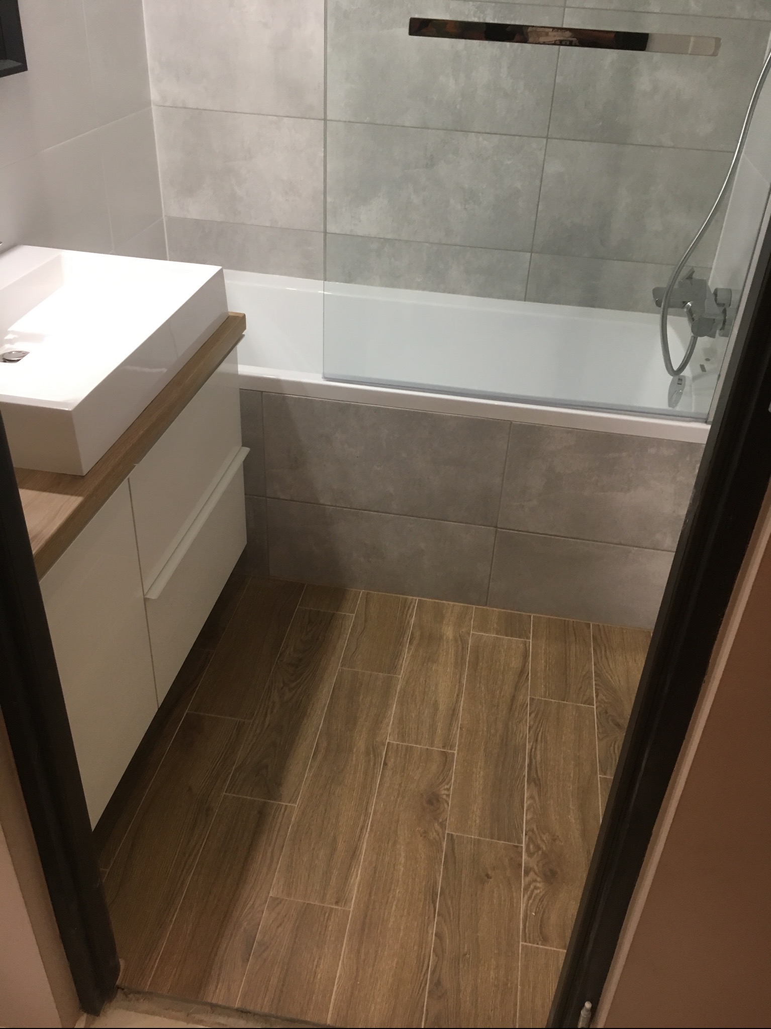 Nowoczesna łazienka z drewnopodobną podłogą, białą umywalką nablatową na szafce z drewnianym blatem i szarą glazurą ścienną, widoczna wanna z parawanem nawannowym.