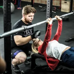 Kobieta leżąca na ławce podnosząca sztangę pod nadzorem mężczyzny w siłowni, z widocznym sprzętem do ćwiczeń i ceglaną ścianą w tle.