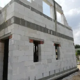 Wznoszenie ścian zewnętrznych domu z białych bloczków, widoczne otwory okienne i drzwiowe z częściowo wmurowaną czerwoną cegłą, ocieplenie fundamentów, w tle zieleń i fragment samochodu dostawczego.