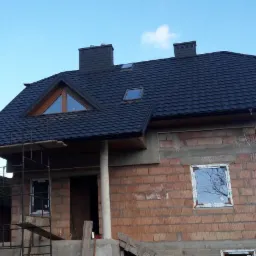 Dom w trakcie budowy z nowym, czarnym dachem z blachodachówki, widoczne rusztowanie, cegły i okna, oraz dwa kominy.