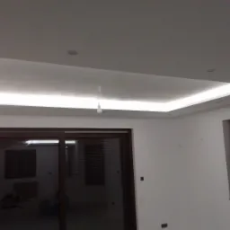 Nowoczesne oświetlenie sufitowe LED z podwieszanym sufitem w jasnym pokoju, widoczna wisząca żarówka na kablu oraz okno z ciemną ramą.