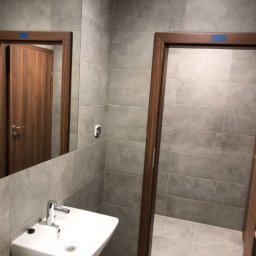 Firma Ogólnobudowlana Kamil Rosochacki - Wykończona łazienka z szarymi płytkami, białą umywalką z baterią, widoczne drewniane ościeżnice drzwi i lustro.