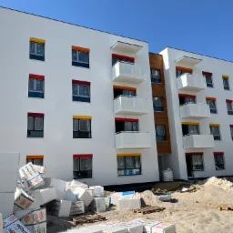 Nowoczesny budynek wielorodzinny z białymi elewacjami i kolorowymi obramowaniami okien (żółte, czerwone, pomarańczowe), widoczne balkony z białymi barierkami, na pierwszym planie stosy białych płyt...