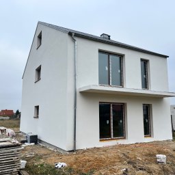 Firma Ogólnobudowlana Kamil Rosochacki - Nowoczesny, dwupiętrowy dom z jasną elewacją, widoczne okna z brązowymi ramami i fragment placu budowy z materiałami budowlanymi.