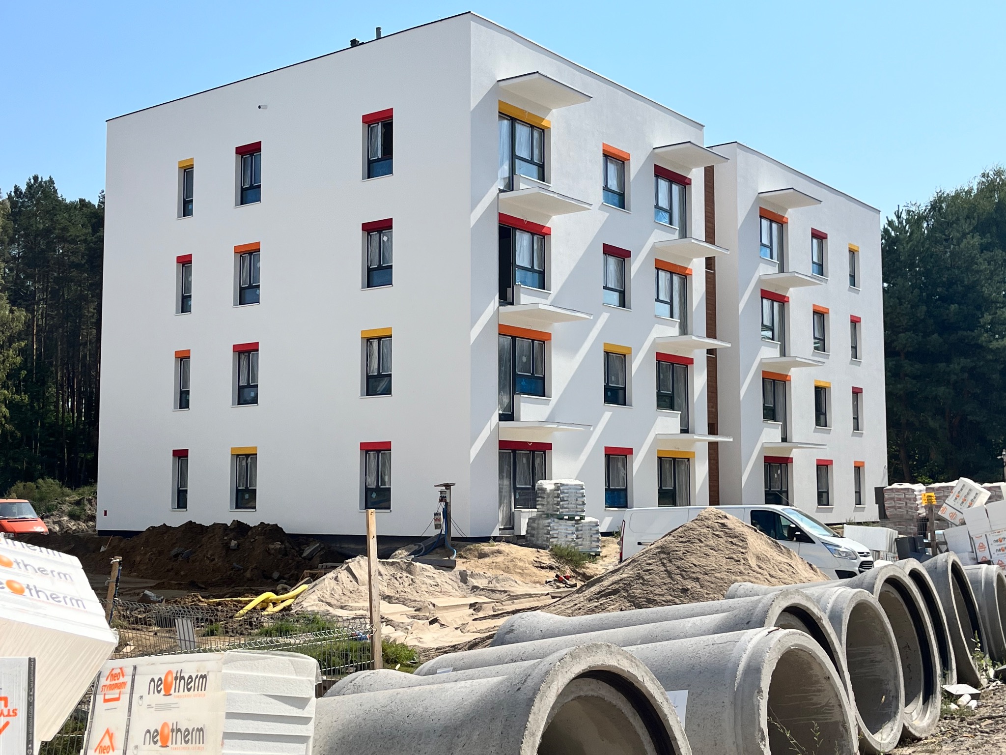 Nowoczesny budynek wielorodzinny z białą elewacją i kolorowymi obramowaniami okien, widoczne materiały budowlane i elementy infrastruktury przed budynkiem.
