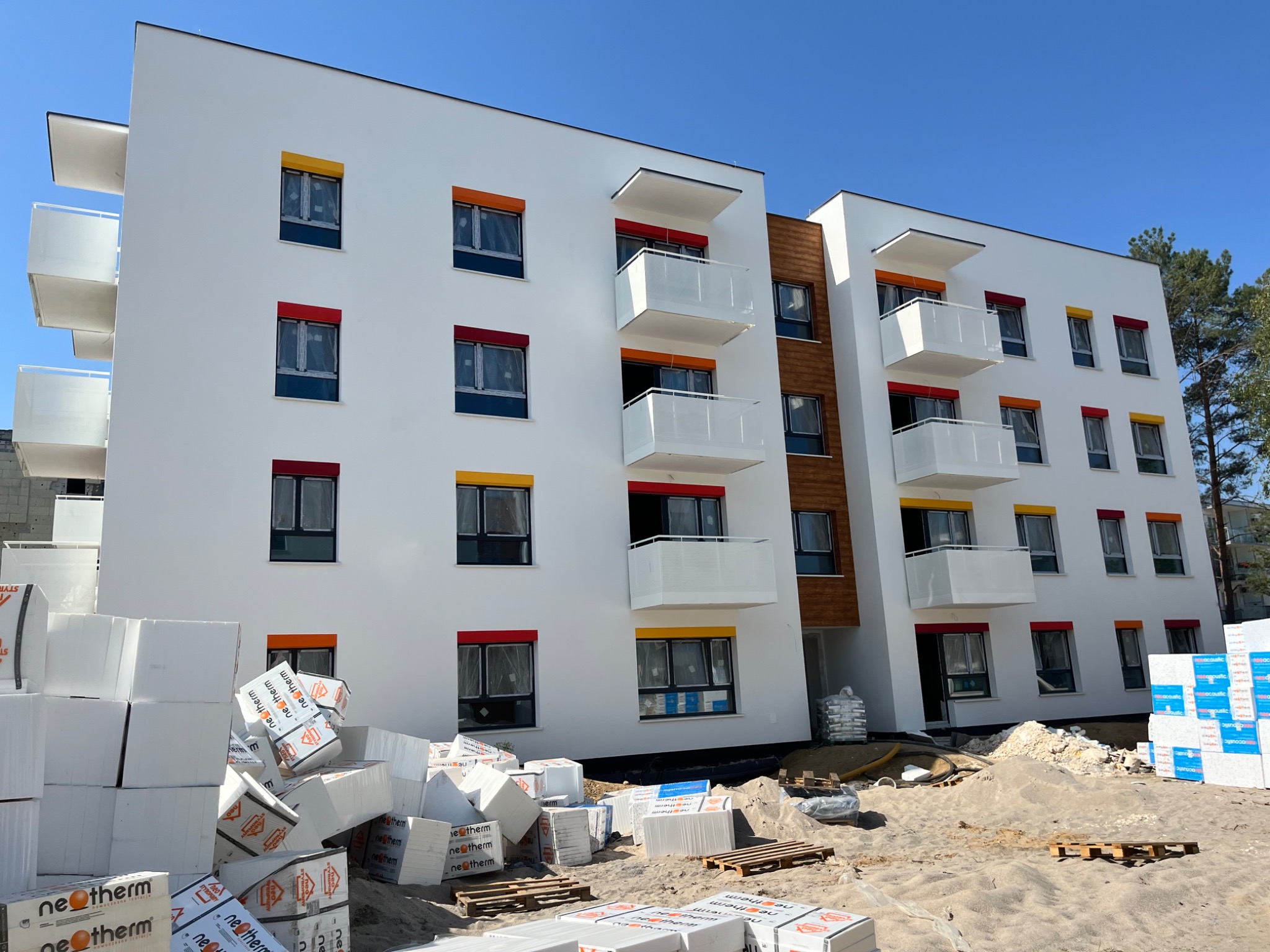 Nowoczesny budynek wielorodzinny z białymi elewacjami i kolorowymi obramowaniami okien (żółte, czerwone, pomarańczowe), widoczne balkony z białymi barierkami, na pierwszym planie stosy białych płyt...