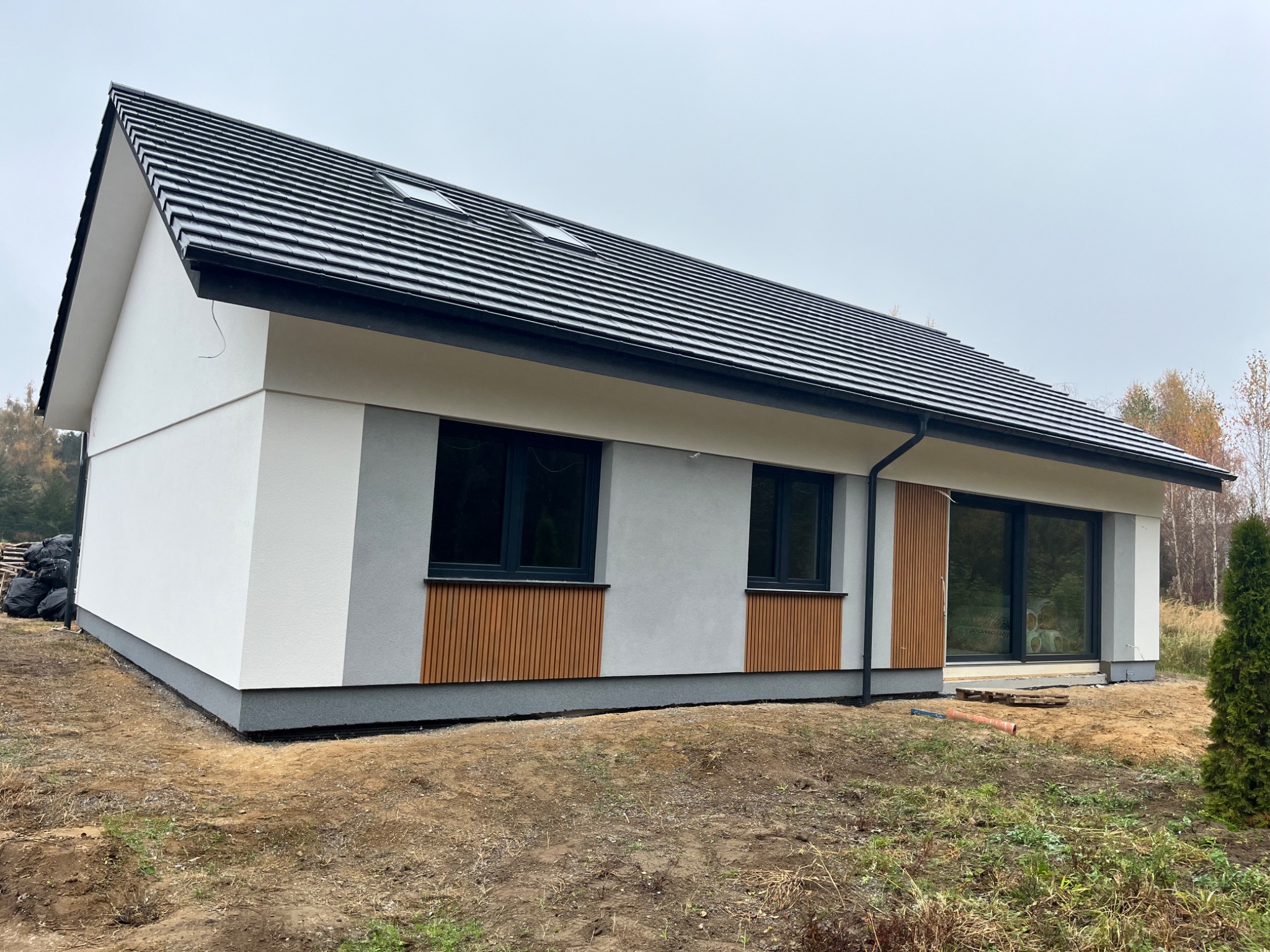 Nowoczesny dom jednorodzinny z jasną elewacją, elementami drewnianymi i ciemną dachówką, widok z zewnątrz na niezagospodarowaną działkę w pochmurny dzień.