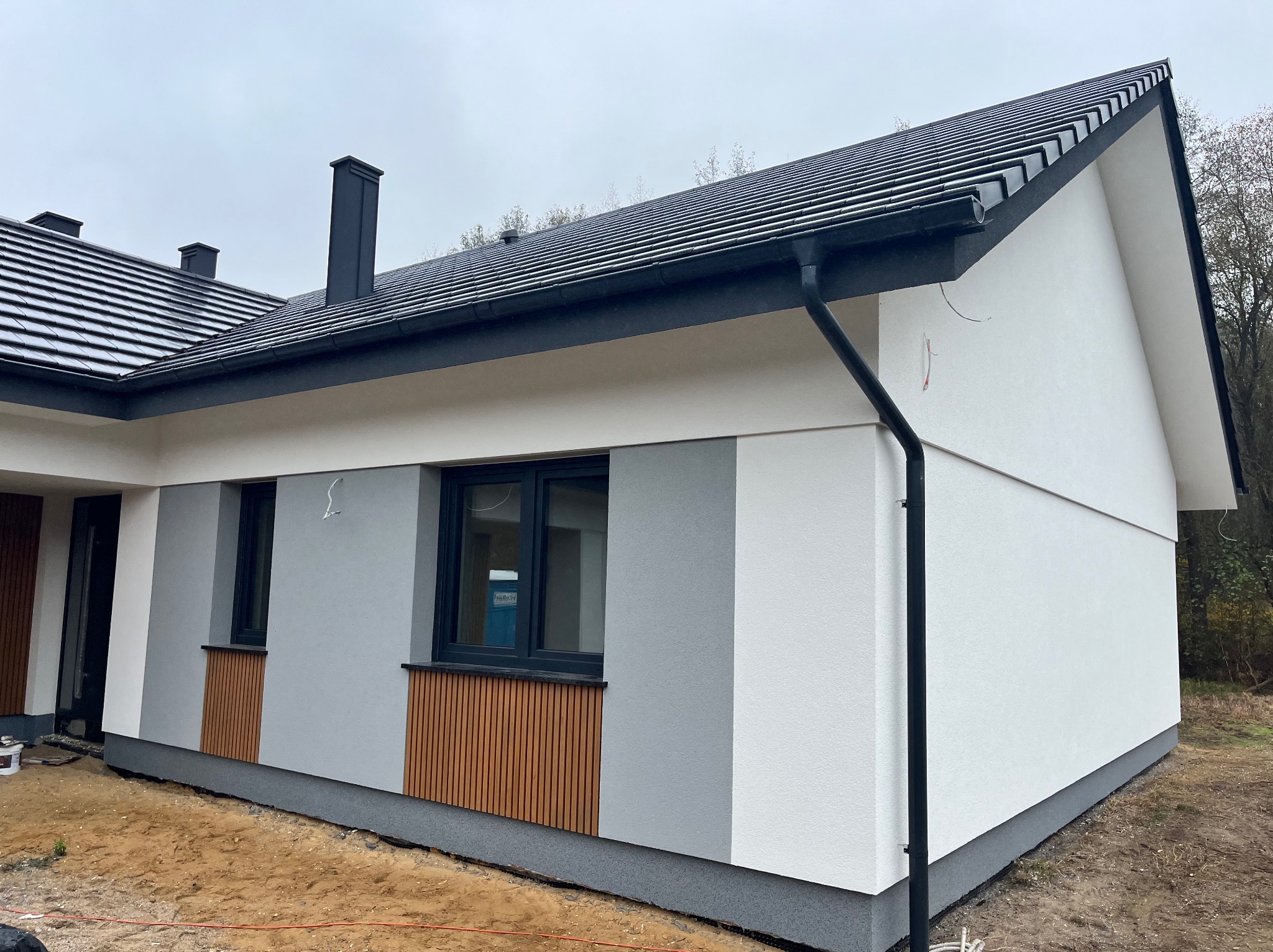 Nowoczesny dom jednorodzinny z elewacją w odcieniach bieli i szarości, z akcentami drewnianymi pod oknami, ciemna dachówka i orynnowanie, widoczne elementy wykończeniowe wokół budynku.