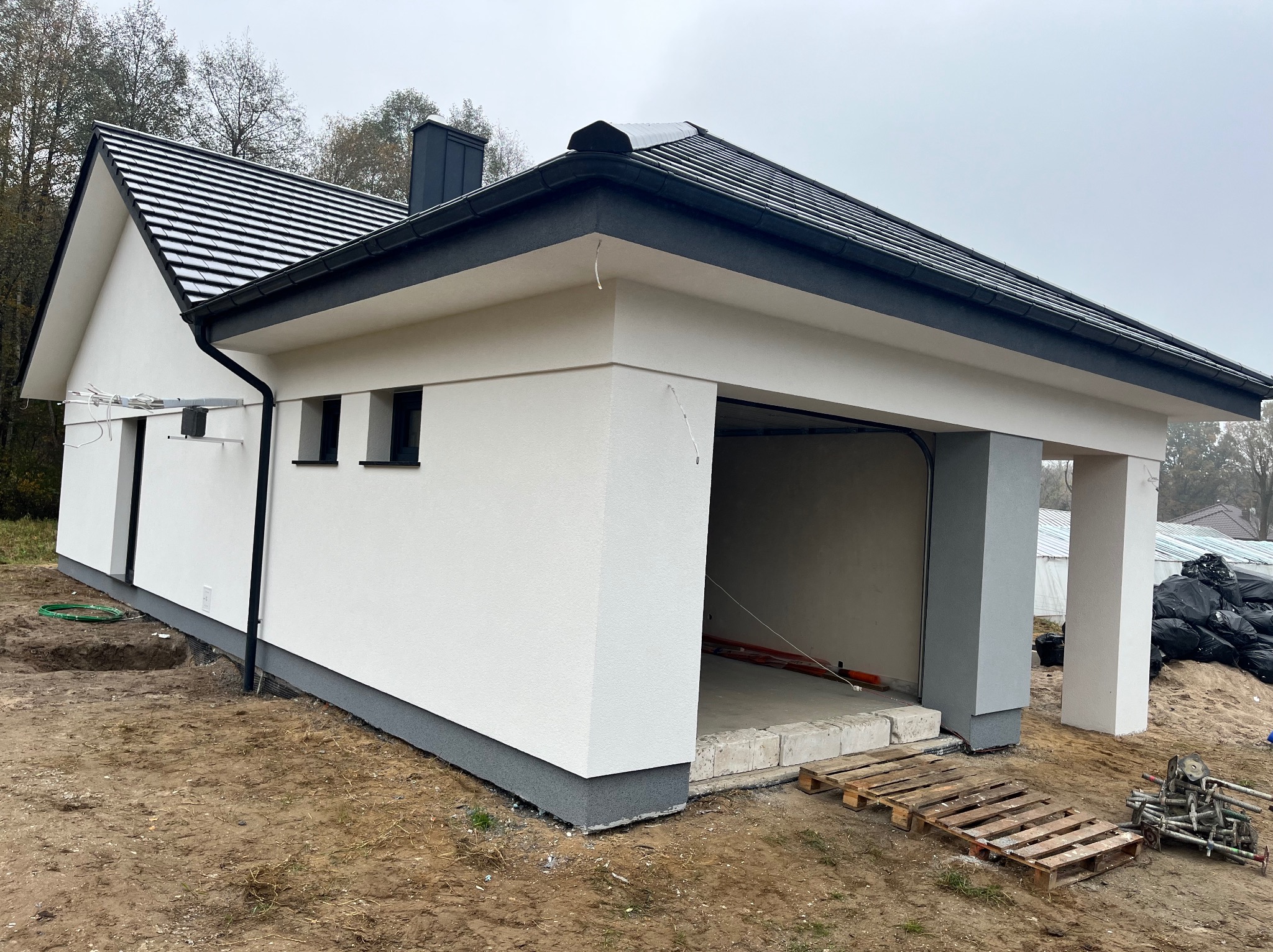 Nowoczesny, biały budynek z garażem w trakcie wykańczania, widoczne elementy konstrukcyjne, palety i narzędzia budowlane na zewnątrz, teren wokół budynku częściowo przygotowany.