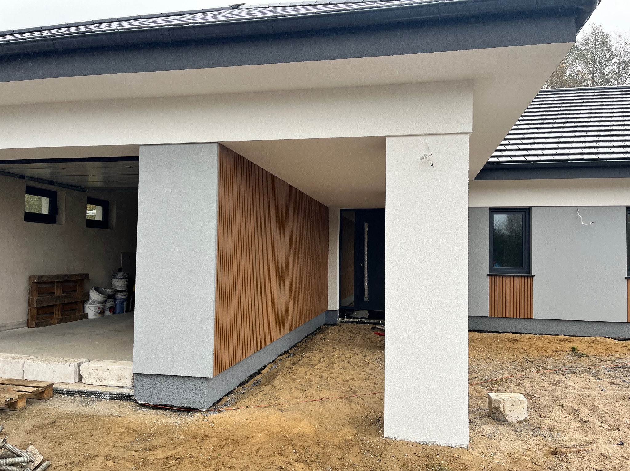Nowoczesny dom jednorodzinny w trakcie wykończenia, widoczna elewacja z jasnoszarego tynku i drewnianych paneli, odkryty fundament i teren wokół domu w stanie surowym, garaż z widocznym wnętrzem.