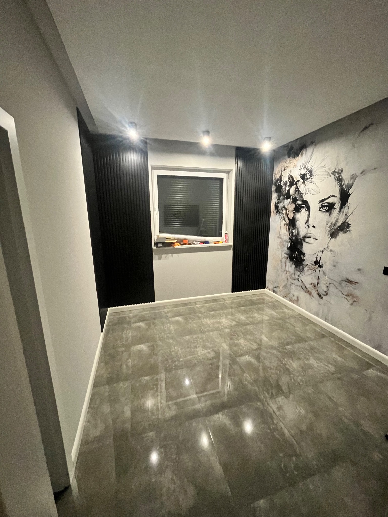 Pusty pokój z szarą, błyszczącą podłogą, białą listwą przypodłogową, ścianą z tapetą przedstawiającą portret kobiety w stylu szkicu, czarnymi pionowymi panelami dekoracyjnymi po bokach okna...