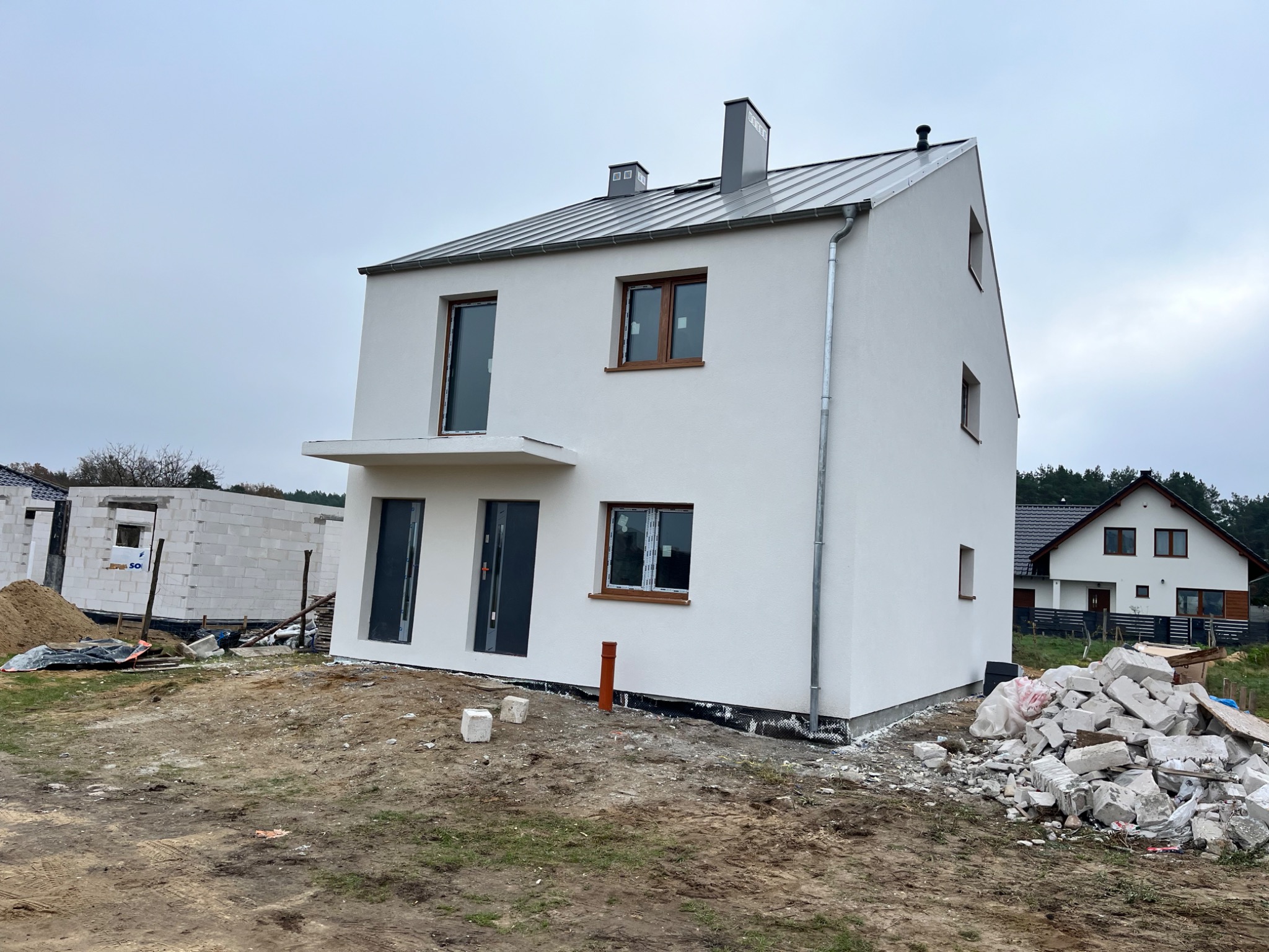 Nowoczesny, dwukondygnacyjny dom z białą elewacją i szarym dachem z blachy, widok z zewnątrz, na pierwszym planie gruz i niedokończona ziemia, w tle inny dom i fragment budynku w trakcie budowy.