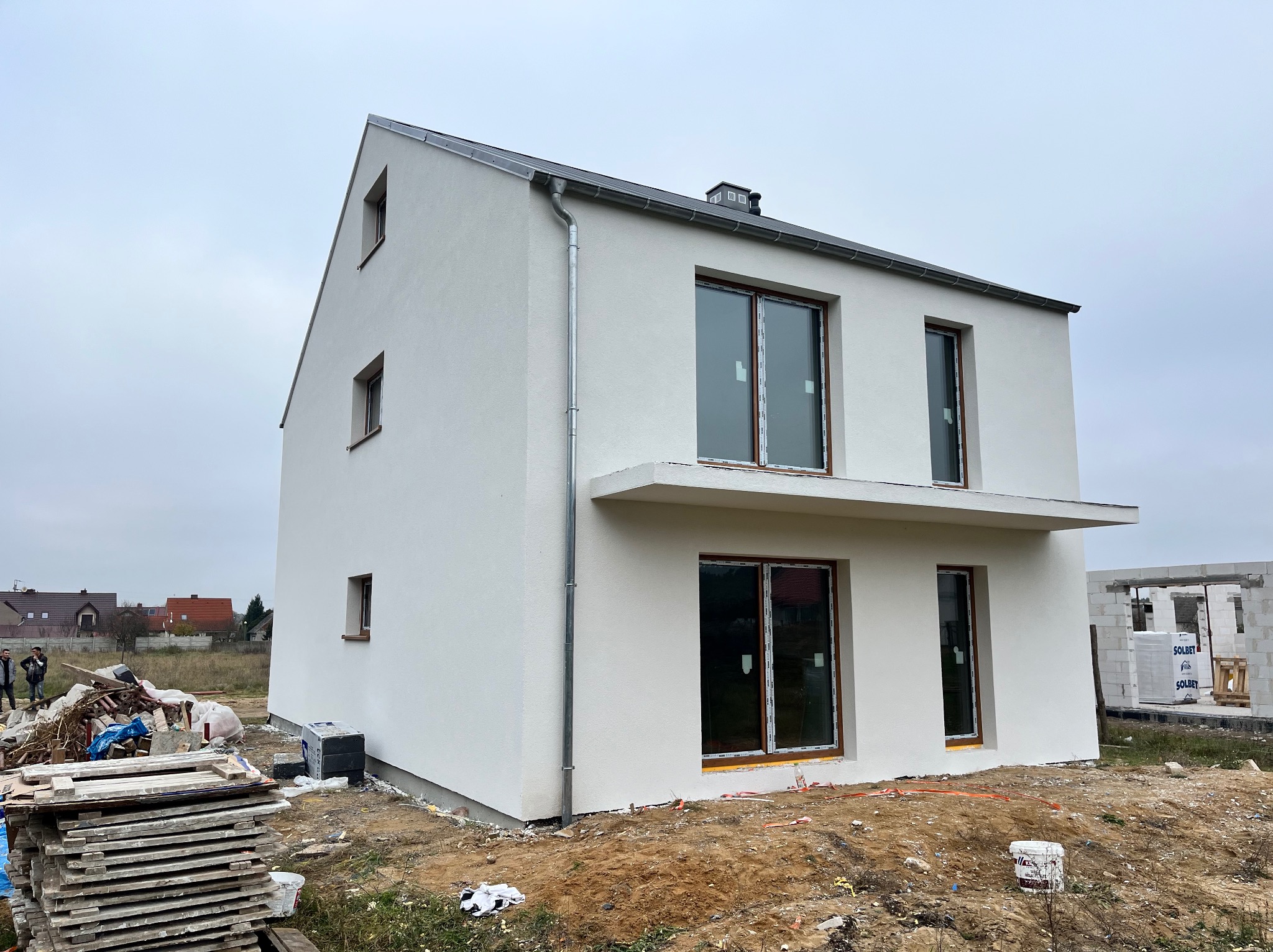 Nowoczesny, dwupiętrowy dom z jasną elewacją, widoczne okna z brązowymi ramami i fragment placu budowy z materiałami budowlanymi.