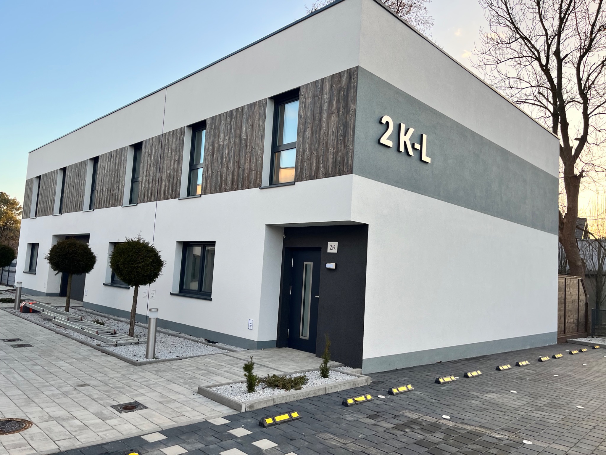Nowoczesny, dwukondygnacyjny budynek z elewacją w odcieniach bieli, szarości i drewna, z numerem 2K-L, otoczony kostką brukową i niską zielenią, drabina leży na ziemi.