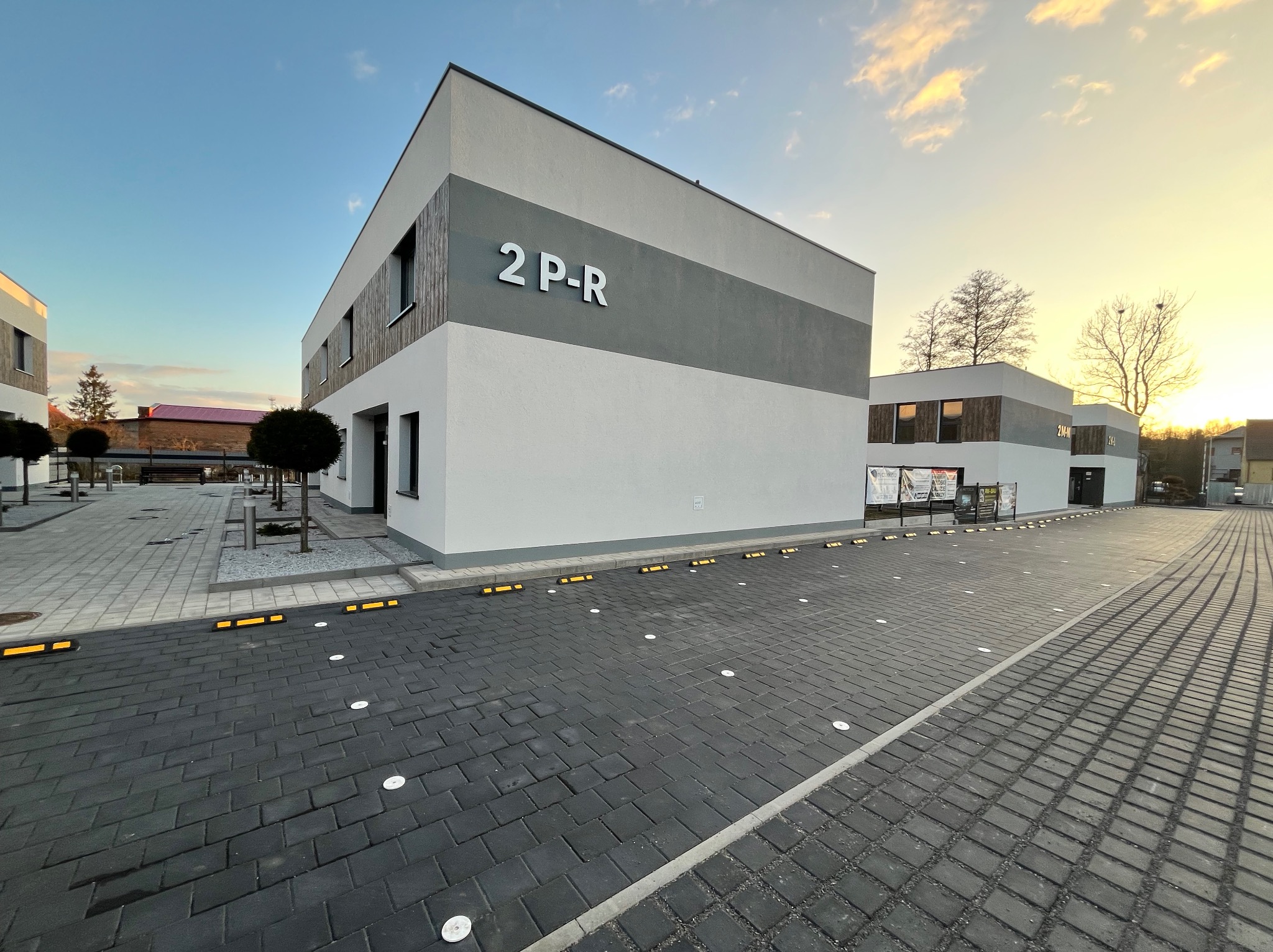 Nowoczesny budynek biurowy z elewacją w odcieniach bieli i szarości, z drewnianymi panelami i dużym napisem '2 P-R', sfotografowany o zachodzie słońca, z widocznym brukowanym parkingiem z żółtymi...