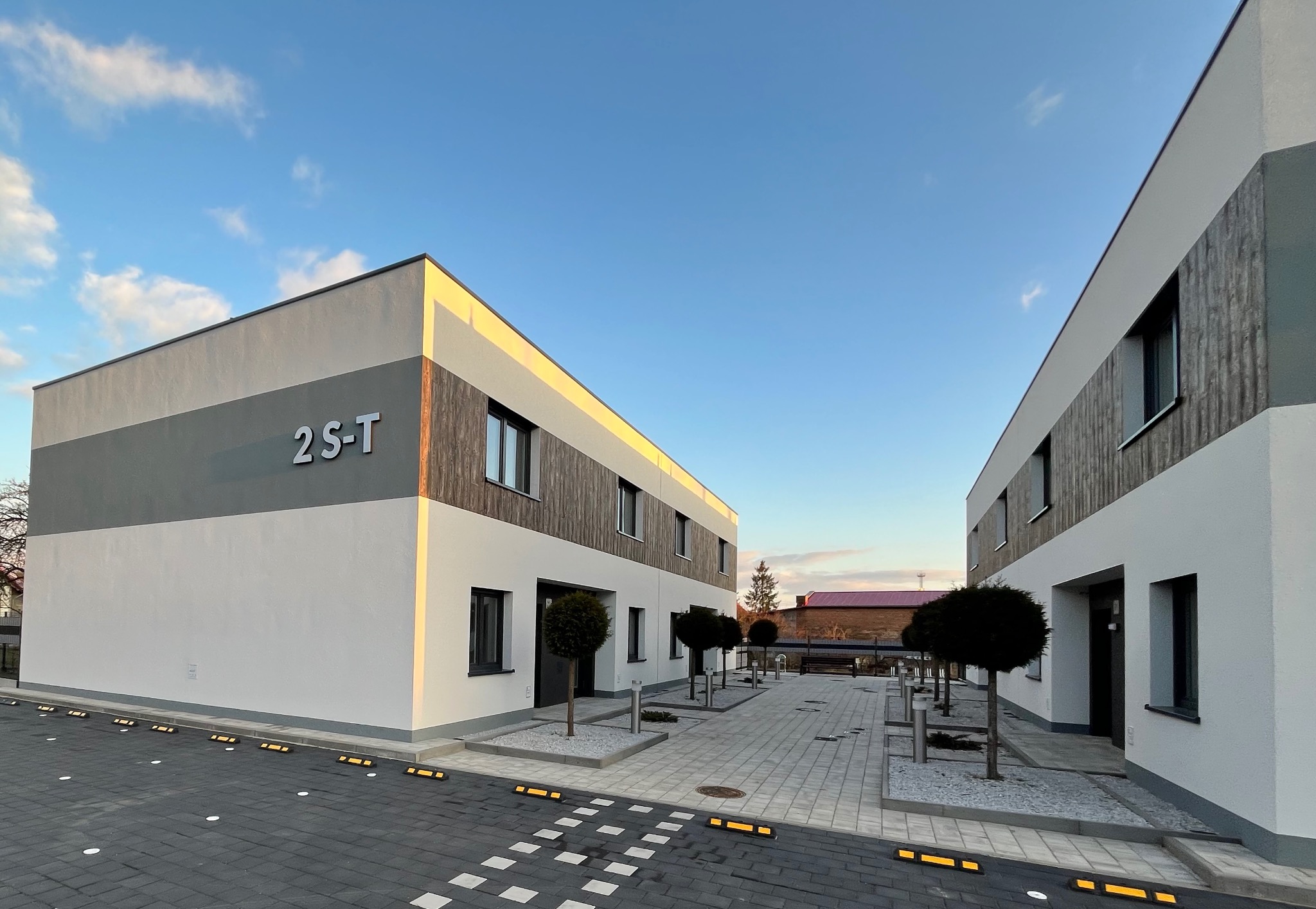 Nowoczesny budynek wielorodzinny z elewacją w odcieniach szarości i bieli, z elementami drewnianymi, oświetlony popołudniowym słońcem, z widocznymi numerami budynków '2S-T' i charakterystycznymi...