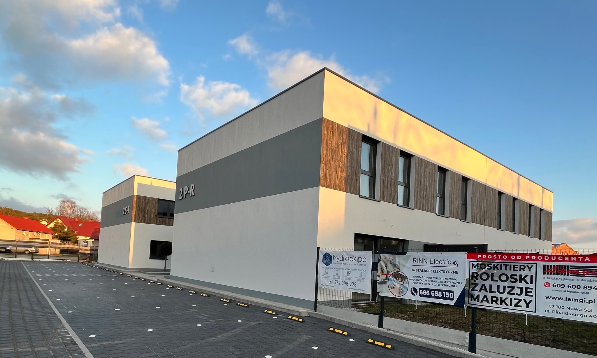 Nowoczesny budynek biurowy z szarą i białą elewacją, akcentami z drewnopodobnej okładziny, widoczne reklamy firm na ogrodzeniu, kostka brukowa na parkingu, błękitne niebo