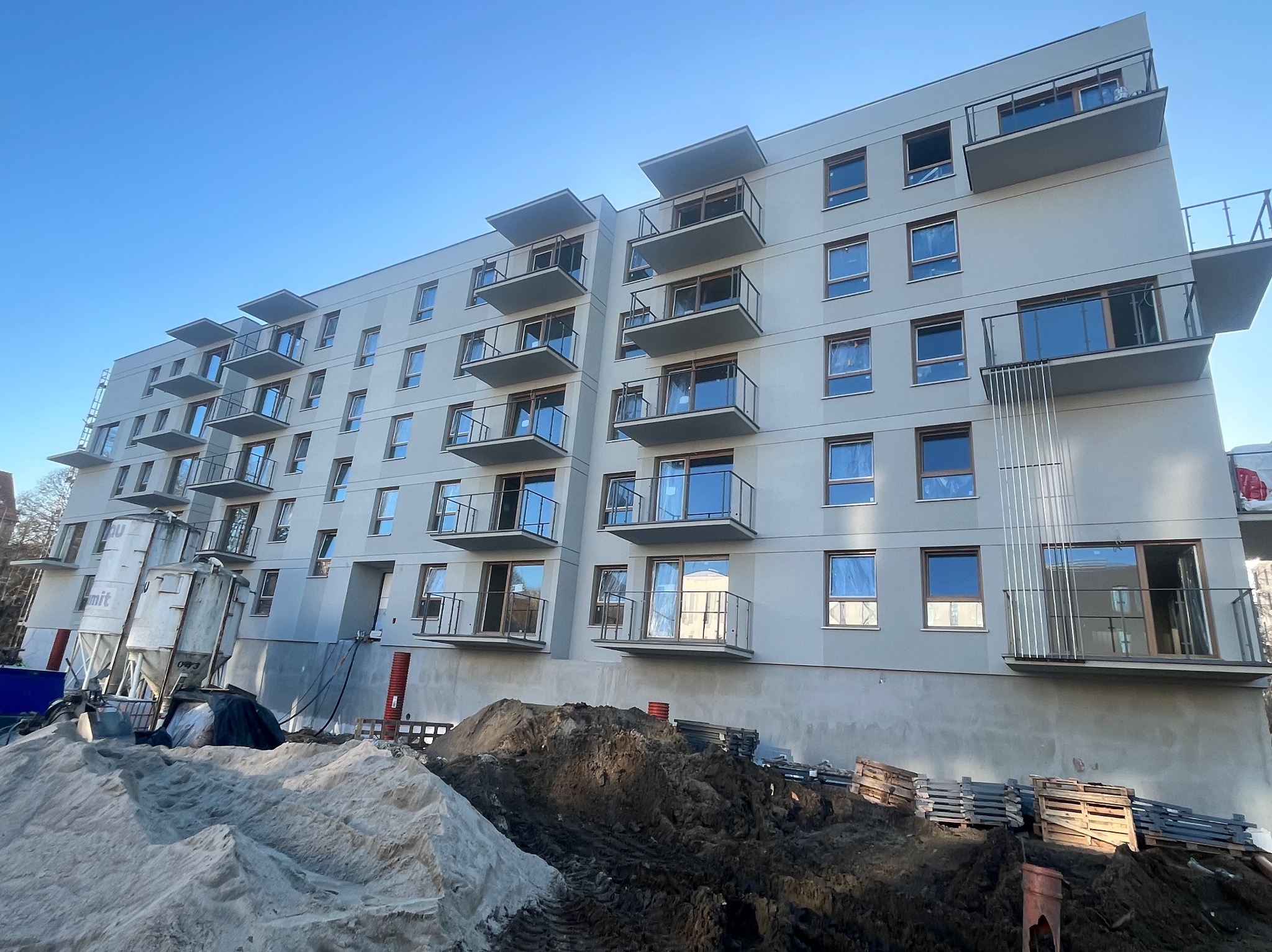 Nowoczesny budynek wielorodzinny z balkonami, w trakcie prac wykończeniowych elewacji. Widoczne rusztowania i materiały budowlane.