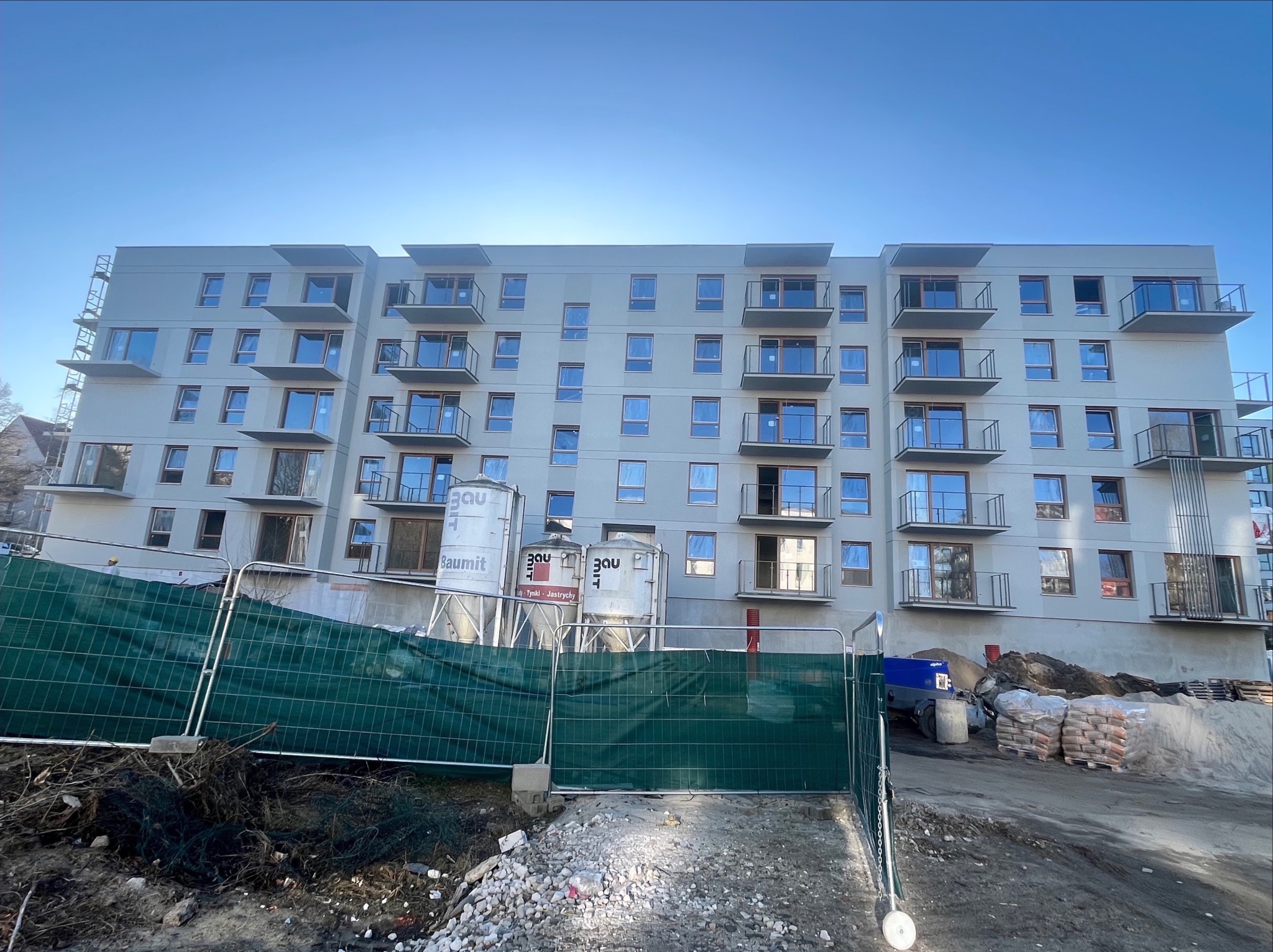 Nowoczesny, wielopiętrowy budynek mieszkalny z balkonami, w trakcie prac wykończeniowych elewacji. Widoczne silosy z zaprawą i materiały budowlane.