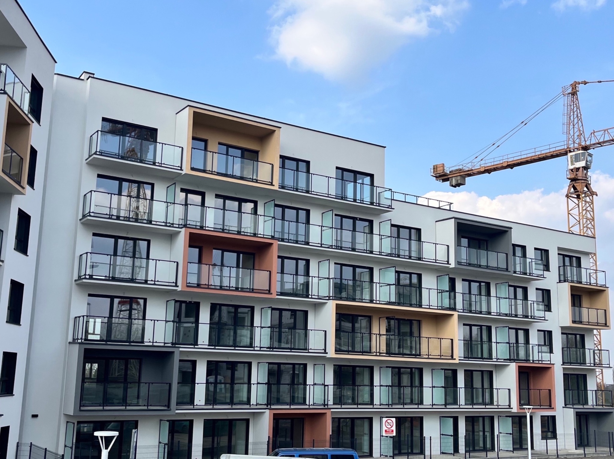Nowoczesny budynek wielorodzinny z balkonami o szklanych balustradach i kolorowych wnękach, w tle żuraw budowlany i błękitne niebo.
