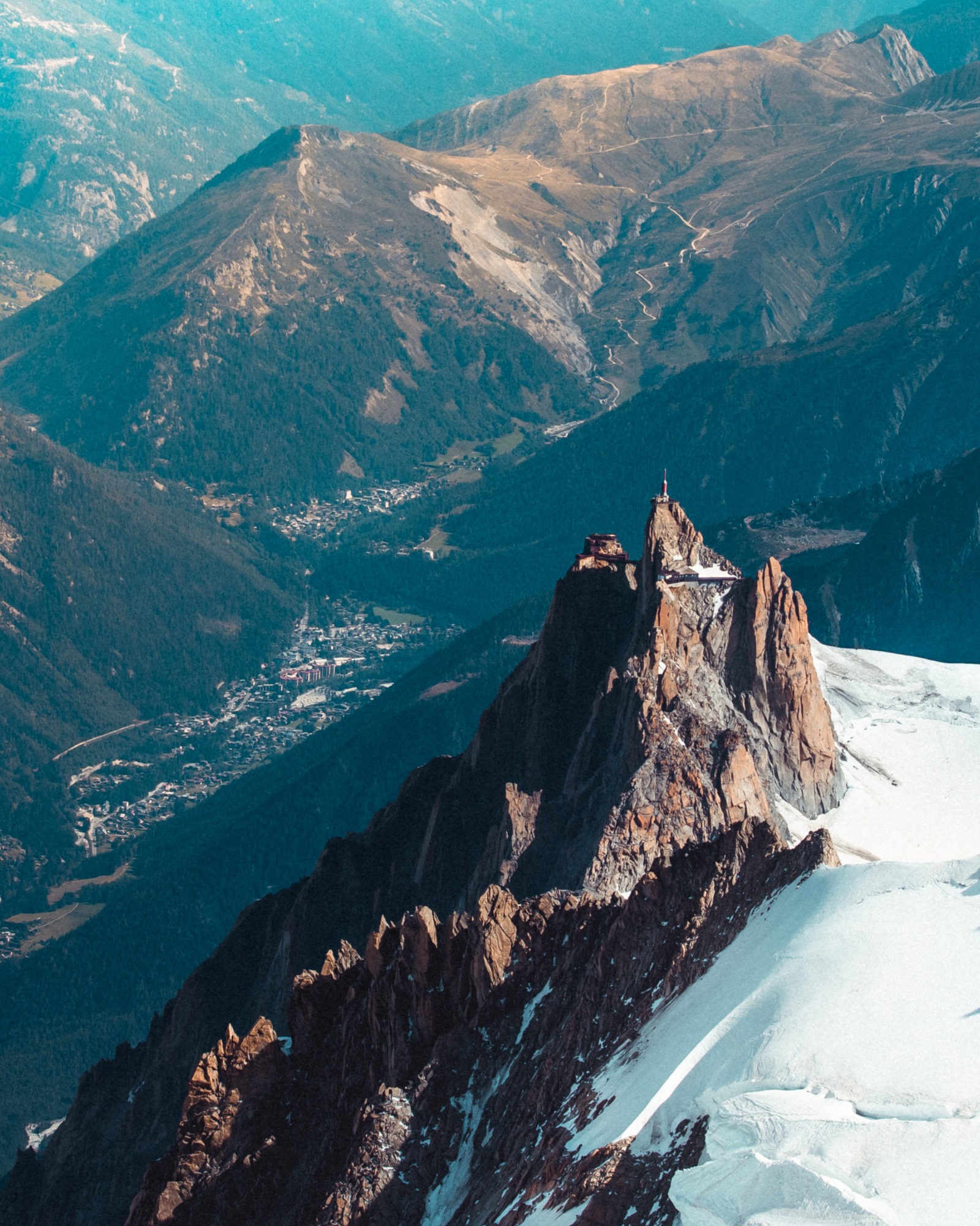 Widok z lotu ptaka na skalisty szczyt z budynkami i anteną, otoczony śnieżnymi polami i zielonymi dolinami z miastem w tle.