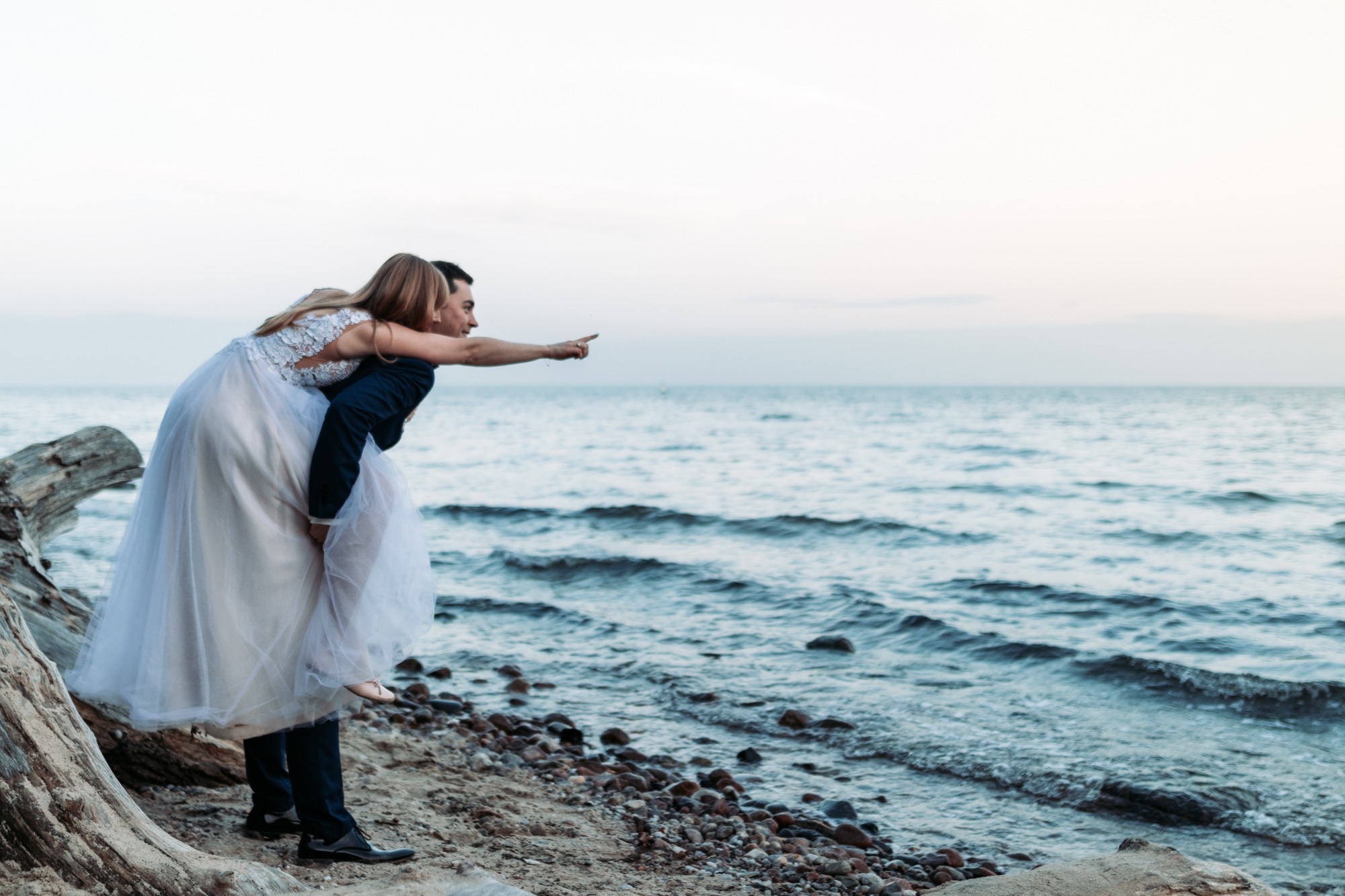 Para młoda w Iławie na plaży: pan młody niesie pannę młodą na barana, oboje wskazują palcem na morze, widoczne kamienie na brzegu i fragment pnia drzewa.