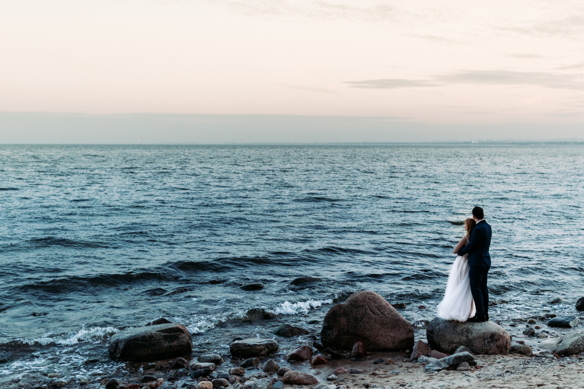 Para młoda, on w granatowym garniturze, ona w białej sukni, obejmują się na kamieniu na plaży, patrząc na morze o zachodzie słońca.