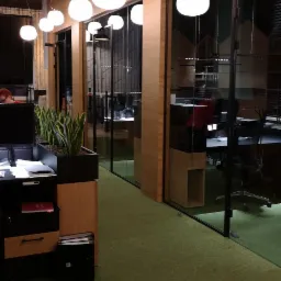 Nowoczesne wnętrze biurowe z szklanymi ścianami, oświetleniem kulistym i zieloną wykładziną dywanową, widok na stanowiska pracy z komputerami.