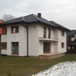 Nowoczesny, dwukondygnacyjny dom z białym tynkiem, ciemną dachówką i drewnianymi ramami okiennymi, otoczony trawnikiem z resztkami śniegu.