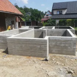 Surowe, betonowe fundamenty domu jednorodzinnego w trakcie budowy, widoczne szalunki i zbrojenia, w tle zabudowa jednorodzinna i zieleń.