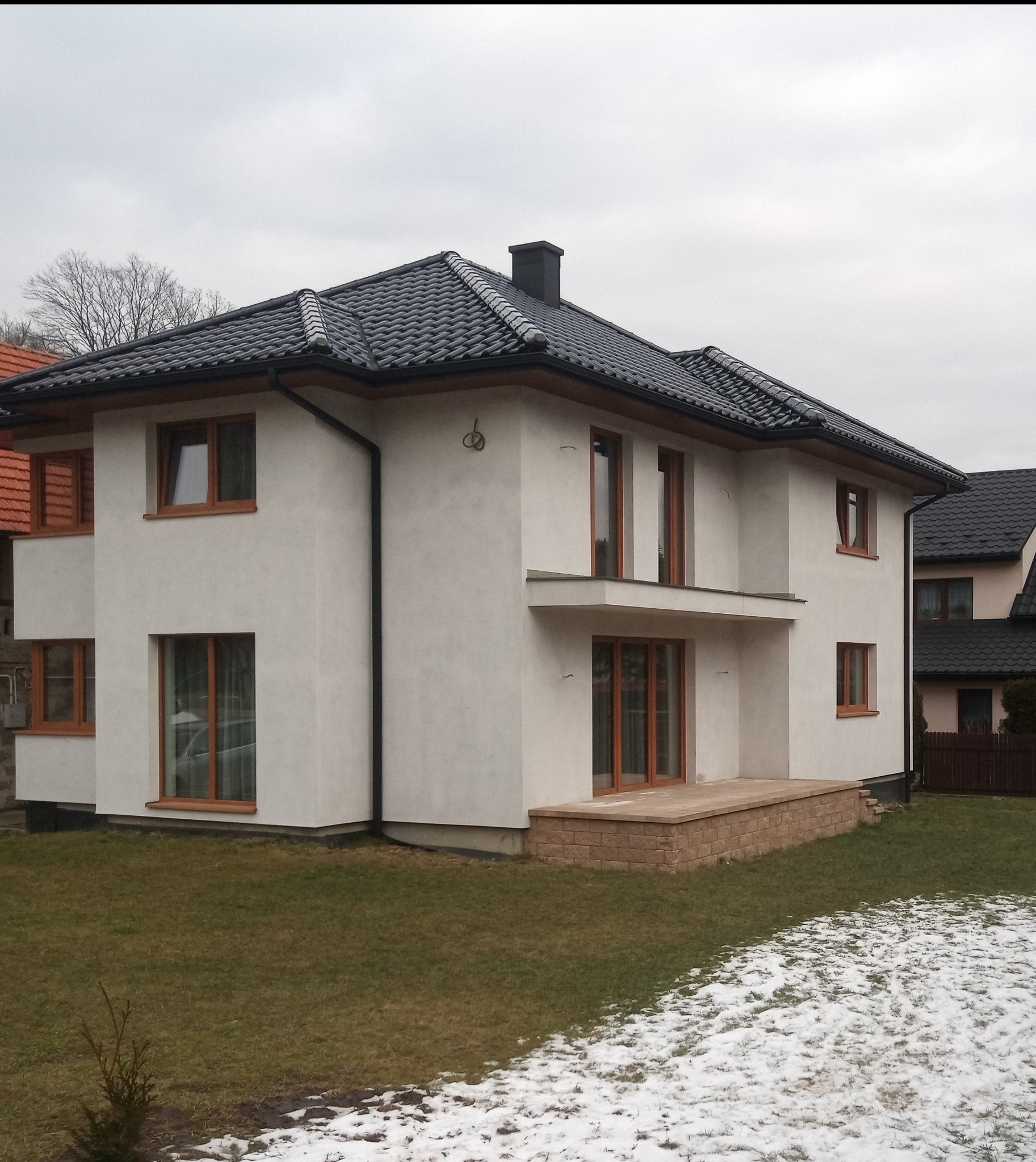 Nowoczesny, dwukondygnacyjny dom z białym tynkiem, ciemną dachówką i drewnianymi ramami okiennymi, otoczony trawnikiem z resztkami śniegu.