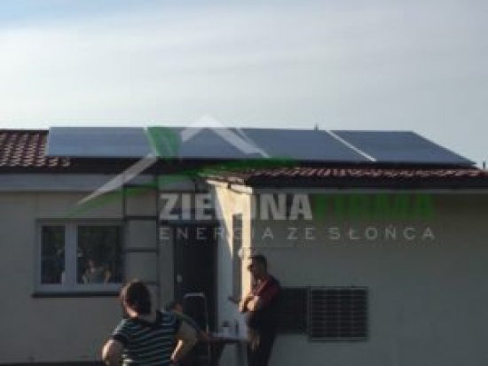 Panele słoneczne na dachu domu jednorodzinnego z widocznym logo firmy 'Zielona Energia ze Słońca' na ścianie budynku, w tle błękitne niebo.