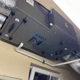 Szara centrala wentylacyjna z rekuperacją, model EBRO NORTADA, zamontowana pod sufitem, widoczne białe rury rozprowadzające powietrze i fragment otwartego okna PCV w tle.