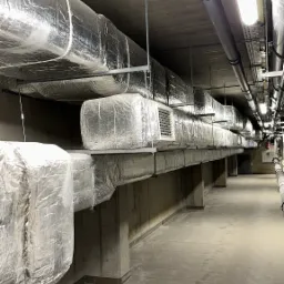 Długi korytarz z systemem wentylacyjnym pod sufitem, rury owinięte srebrną folią izolacyjną, betonowe filary podtrzymujące instalację, płytki podłogowe w odcieniach szarości.