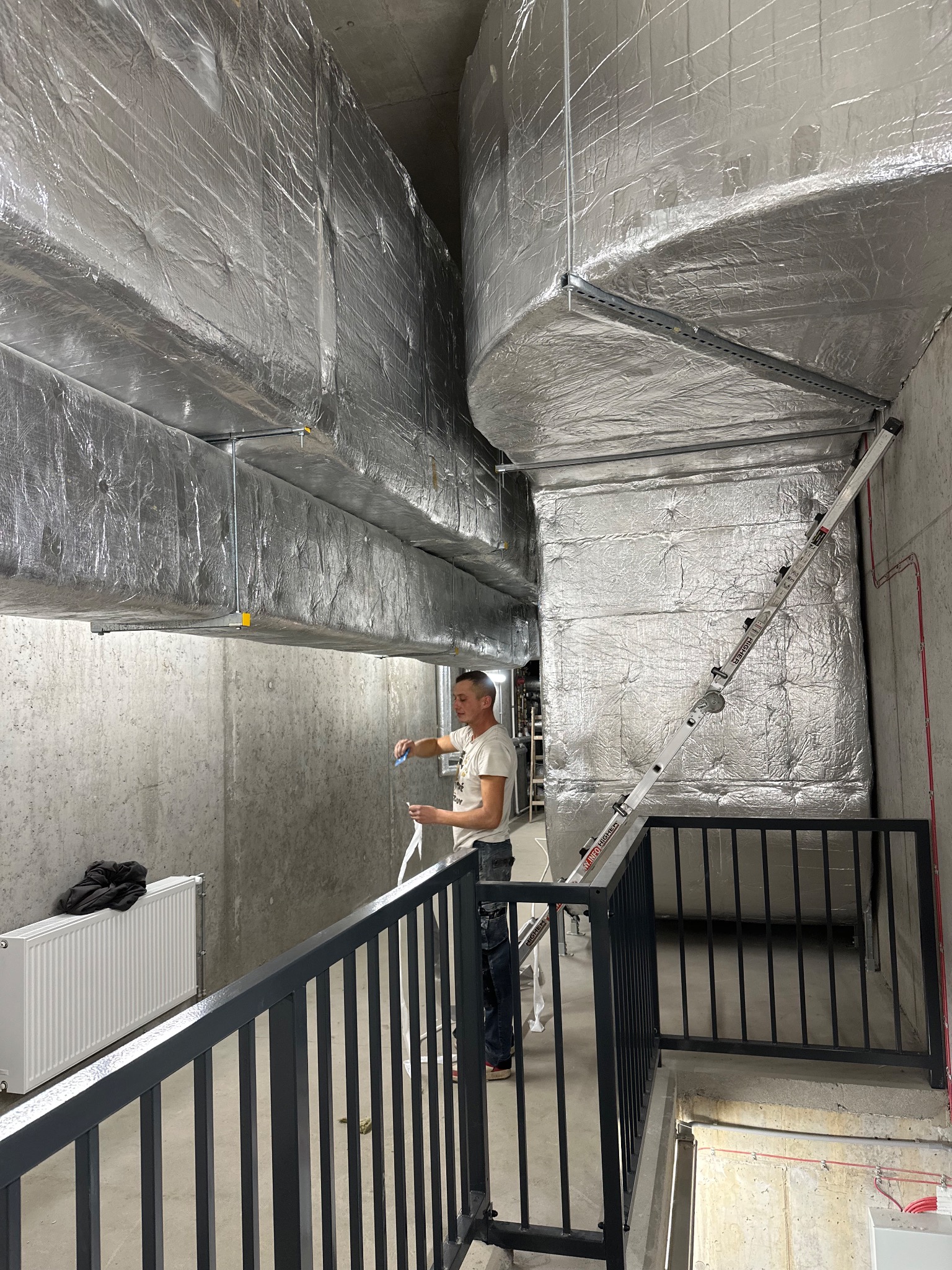 Instalator mierzy taśmą izolacyjną owinięty kanał wentylacyjny w piwnicy, drabina oparta o ścianę, widoczna balustrada schodów i grzejnik.