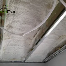 Poddasze ocieplone białą pianką PUR, widoczne krokwie drewniane, profile metalowe, instalacja elektryczna w peszlu oraz logo firmy GreenPur w lewym górnym rogu.