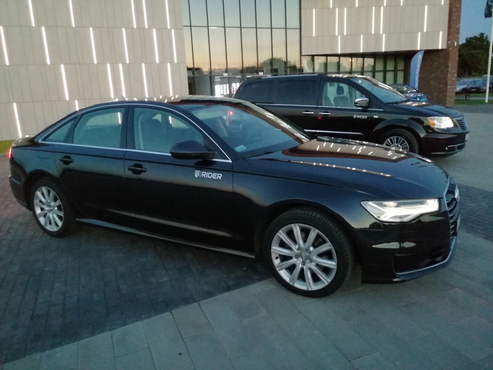 Dwa czarne samochody, sedan Audi i minivan Chrysler, zaparkowane przed nowoczesnym budynkiem z oświetleniem liniowym. Na drzwiach sedana widoczne logo firmy RIDER.